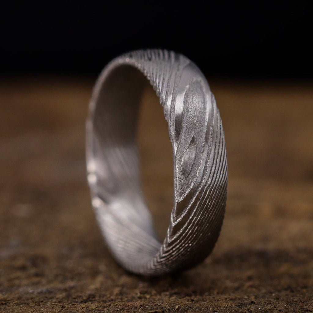 A Damascus Stainless Steel wedding ring with woodgrain effect pattern sits on top of a wooden surface.