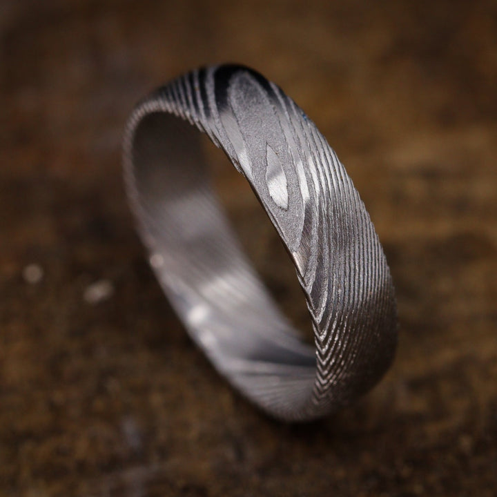 A Damascus Stainless Steel wedding ring with woodgrain effect pattern sits on top of a wooden surface.