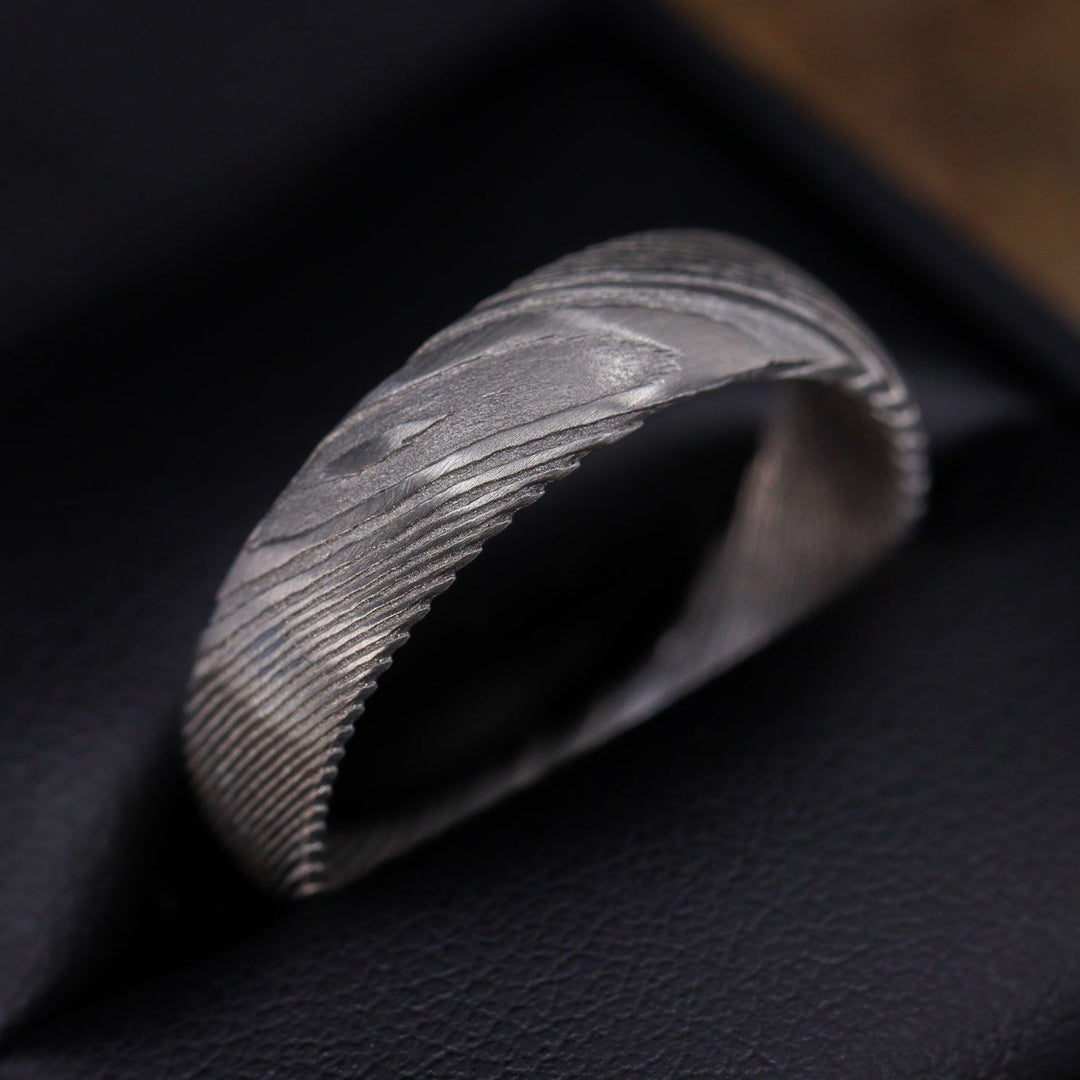 A Damascus Stainless Steel wedding ring with woodgrain effect pattern sits on top of a wooden surface.