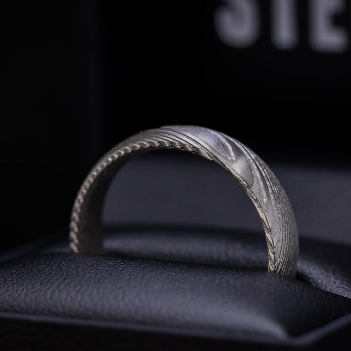 A Damascus Stainless Steel wedding ring with woodgrain effect pattern sits on top of a wooden surface.